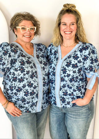 light blue top with navy flowers and red centers - light blue band around v-neck and straight down the front with blue and white striped trim along side it - light blue and striped bow at cuff
