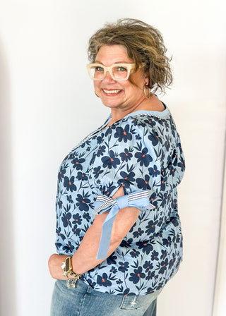light blue top with navy flowers and red centers - light blue band around v-neck and straight down the front with blue and white striped trim along side it - light blue and striped bow at cuff