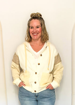 A smiling woman with blonde hair in a bun stands against a plain white wall wearing a cream and beige color-blocked hoodie with brown buttons and blue jeans.