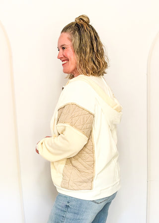A woman wearing a cream and beige color-blocked hoodie with quilted sleeves, standing against a white wall.
