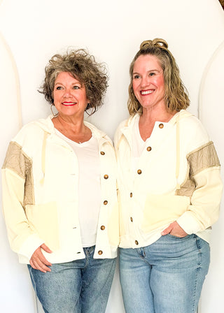 Two women standing side-by-side wearing matching cream-colored hooded button-up jackets with beige sleeve panels and blue jeans, set against a white arched studio backdrop.