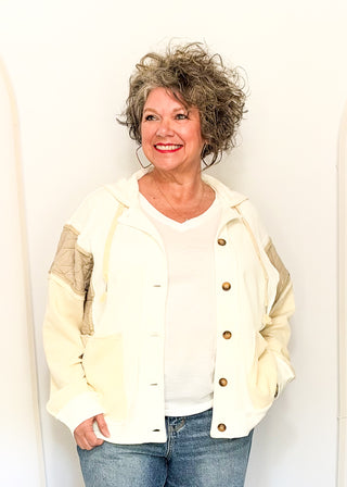 A smiling woman with curly gray hair wearing a cream-colored hooded jacket with brown buttons and textured sleeves, paired with a white top and blue jeans, standing against a plain white wall.