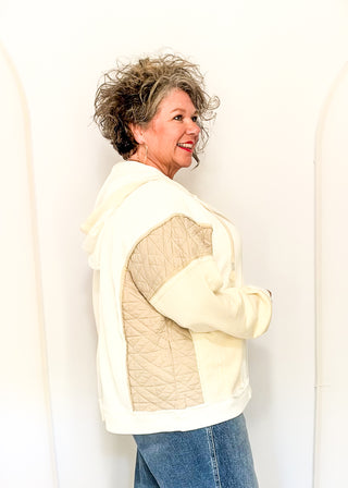 A woman wearing a cream-colored hooded jacket with quilted beige shoulder panels, paired with blue jeans, standing against a white arched wall.