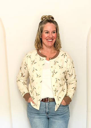A smiling woman with blonde hair in a bun stands against a white wall, wearing a cream-colored bomber jacket with a brown and black bird pattern, brown ribbed trim, and gold buttons, paired with a white top and blue jeans.