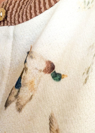 Close-up of a cream-colored knit garment featuring a delicate, hand-painted style bird pattern in muted earthy tones of brown, green, and blue, with a brown striped collar detail at the top.