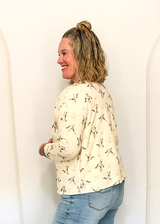 A woman wearing a cream-colored long-sleeve top with a black bird print, paired with blue jeans, standing against a white wall.