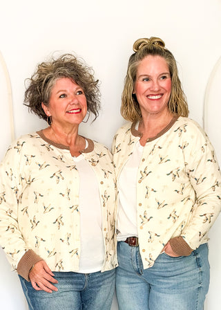 Two smiling women standing side by side wearing matching cream-colored button-up cardigans with a brown bird pattern, white shirts, and blue jeans against a white arched wall.