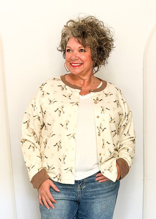 A woman with curly gray hair wearing a cream bomber jacket with a brown and black dragonfly pattern, a white t-shirt, and blue jeans, standing against a white wall.