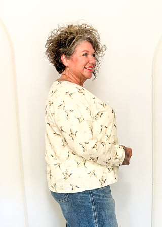 A woman with short, curly gray hair is shown from the side and back, wearing a cream-colored long-sleeve top with a small brown and black bird pattern, paired with blue jeans.
