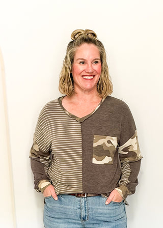 A smiling woman with blonde hair in a bun wears a long-sleeve V-neck top with a color-blocked design: beige and brown horizontal stripes on one side, solid brown on the other, and a camouflage print pocket on the front. She is wearing blue jeans against a plain white wall.