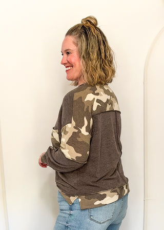 A woman wearing a brown long-sleeve top with camouflage-patterned shoulder and sleeve panels, paired with blue jeans, standing against a plain white wall.