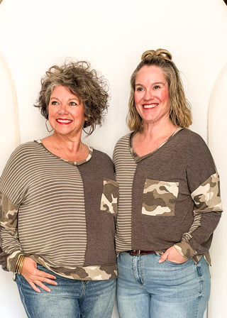 Two women modeling matching long-sleeve V-neck tops with a color-blocked design featuring horizontal stripes on one side and a camouflage pattern on the other, paired with blue jeans against a white arched wall.