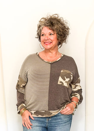 A woman with curly gray hair wearing a brown and beige color-blocked long-sleeve top with a camouflage pocket and striped pattern, paired with blue jeans, standing against a white wall.