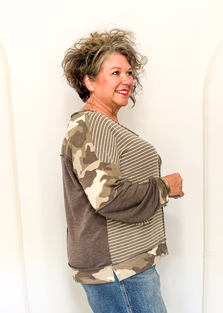 A woman modeling a long-sleeve top with a mixed pattern design, featuring solid brown on one side and camouflage on the other, paired with blue jeans against a plain white background.
