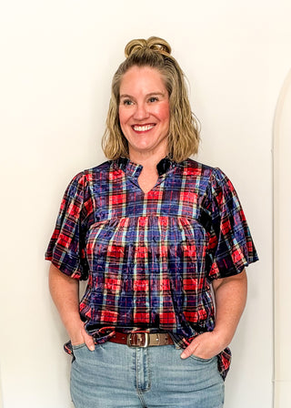A smiling woman with blonde hair in a bun wears a red, navy, and white plaid short-sleeve blouse tucked into light-wash blue jeans with a brown belt, standing against a plain off-white wall.