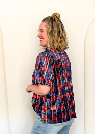 A woman with blonde hair in a high ponytail is shown from a side-back angle, wearing a red, navy, and blue plaid short-sleeve button-up shirt and light wash jeans against a plain off-white wall.