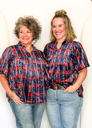 Two women standing side by side wearing matching short-sleeve plaid button-up shirts in red, navy, and blue tones, paired with light-wash jeans, against a white arched studio backdrop.