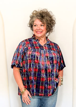 A smiling woman with curly gray hair wearing a red, navy, and white plaid short-sleeve button-up shirt and blue jeans, standing against a plain white wall.