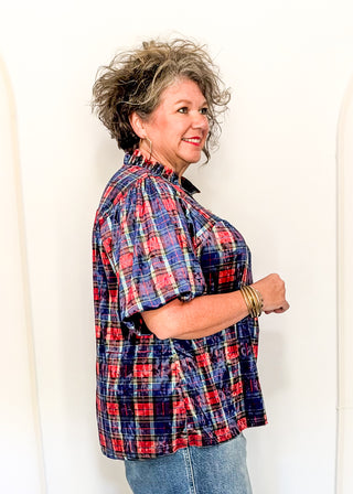 A woman with curly gray hair is shown from the side, wearing a short-sleeve plaid button-up shirt in red, blue, and white, paired with blue jeans, against a plain white background.