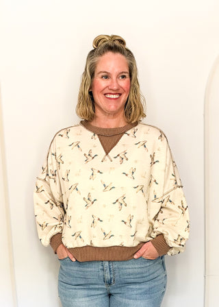 A woman with blonde hair in a bun wearing a cream-colored long-sleeve sweatshirt with a brown ribbed crew neck, cuffs, and hem, paired with blue jeans, standing against a white wall.