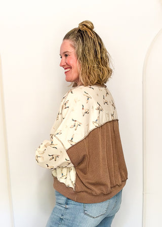 A woman wearing a color-blocked bomber jacket with a cream and brown bird print on one side and a solid brown corduroy back panel, paired with light blue jeans, standing against a white wall.