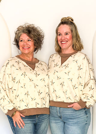 Two women standing side by side wearing matching cream-colored long-sleeve sweaters with a brown ribbed crew neck, hem, and cuffs, featuring a small bird print. They are also wearing blue jeans, set against a plain white wall.