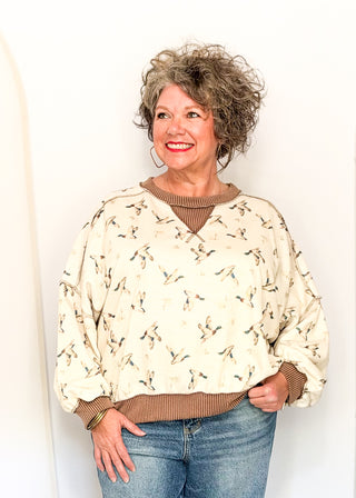 A smiling woman with curly gray hair wearing a cream-colored long-sleeve sweater with a brown and black bird pattern, layered over a brown ribbed knit top and paired with blue jeans, standing against a plain white wall.