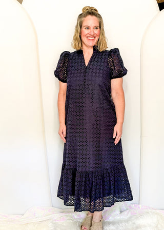 A woman modeling a dark navy blue eyelet midi dress with puff sleeves and a ruffled hem, standing against a white arched backdrop.