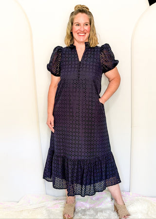 A woman modeling a dark navy midi dress with a black eyelet lace pattern, puff sleeves, and a ruffled hem, standing against a white arched backdrop.