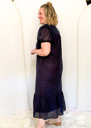 A woman modeling a dark navy blue crochet midi dress with short puff sleeves and a ruffled hem, standing in front of a white arched backdrop.