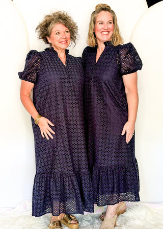 Two women modeling matching dark purple eyelet midi dresses with puff sleeves and ruffled hems, standing against a white studio backdrop.
