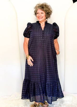 A woman modeling a dark navy blue eyelet midi dress with puff sleeves and a ruffled hem, standing against a white arched backdrop.