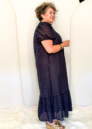 A woman wearing a navy blue short-sleeve midi dress with a textured, dotted eyelet pattern and a ruffled hem, standing in front of a white arched backdrop.