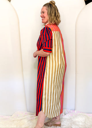 A woman wearing a colorful striped midi dress with red and navy vertical stripes on one side and beige and green vertical stripes on the other, standing in front of a white arched backdrop.