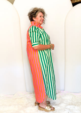 A woman modeling a colorful, long-sleeved maxi dress with vertical stripes. The dress features three distinct color-blocked panels: solid bright orange on the left, green and white stripes on the right, and a solid orange collar.