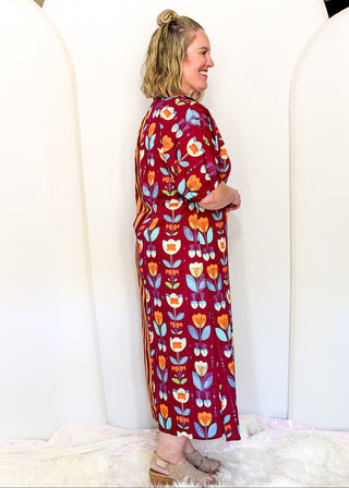 A woman wearing a long-sleeved maxi dress with a retro floral pattern in red, orange, yellow, and blue, standing in front of a white arched backdrop.