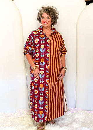 A woman modeling a long collared shirt dress with a split design: one side features a red floral pattern with orange, yellow, and blue flowers, and the other side has vertical orange and brown stripes. She is standing in front of a white arched backdrop.