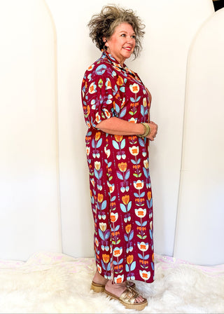 A woman modeling a long-sleeved maxi dress with a retro floral pattern in burgundy, orange, yellow, and light blue, standing against a white arched backdrop.