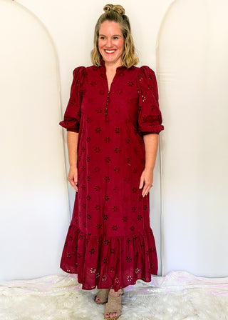 A woman modeling a long-sleeved, midi-length red eyelet dress with a tiered ruffle hem, standing against a white arched backdrop.