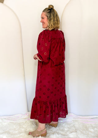 A woman wearing a long-sleeved, deep red eyelet maxi dress with embroidered floral details, standing in front of a white arched backdrop.
