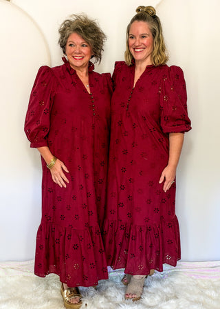 Two women modeling matching burgundy eyelet midi dresses with puff sleeves and tiered ruffles, standing against a white arched wall.