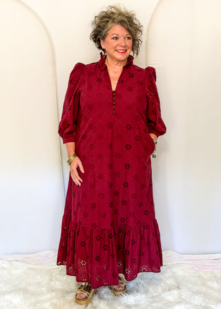 A woman modeling a long-sleeved burgundy eyelet maxi dress with puff sleeves, a buttoned front, and a tiered ruffle hem, standing against a white arched backdrop.