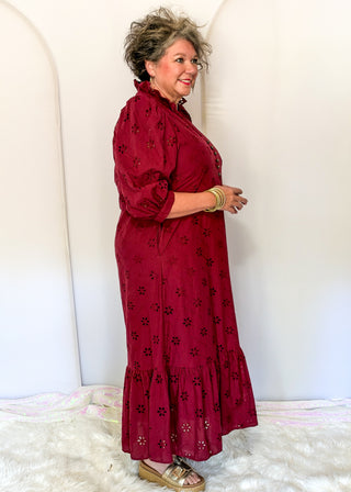 A woman modeling a long-sleeved, high-neck maxi dress in deep burgundy with black floral eyelet embroidery, standing against a white arched backdrop.