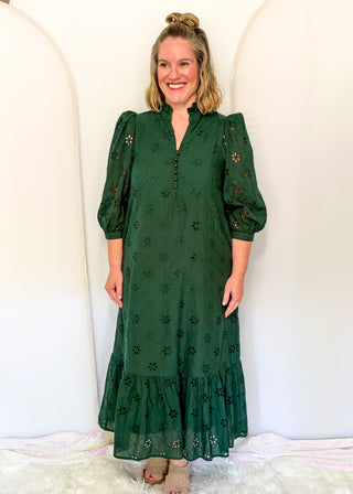 A woman modeling a dark green eyelet midi dress with puff sleeves, a split neckline, and a tiered ruffle hem, standing against a white arched backdrop.