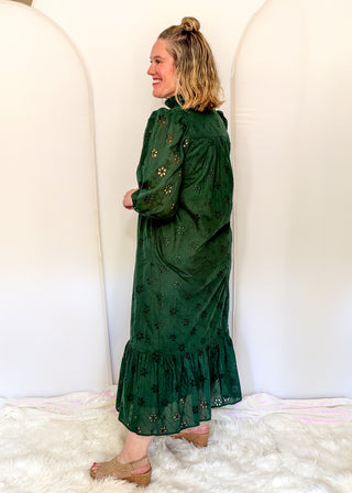 A woman modeling a dark green eyelet midi dress with floral embroidery, standing in front of a white arched backdrop on a white faux fur rug.