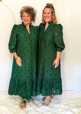 Two women standing side by side wearing matching dark green eyelet midi dresses with puff sleeves, a buttoned front, and a ruffled hem, posed against a white studio backdrop on a white faux-fur surface.
