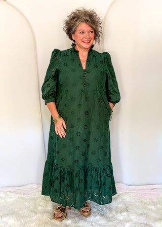 A woman modeling a dark green midi dress with black floral embroidery, puff sleeves, and a tiered ruffle hem, standing against a white arched backdrop.
