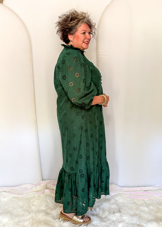 A woman modeling a dark green eyelet maxi dress with floral cutout patterns, standing against a white arched backdrop.