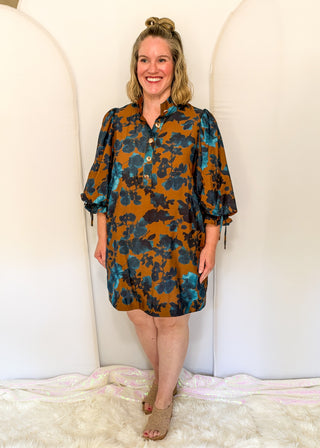 A woman modeling a short-sleeve button-front dress with a brown, teal, and black floral pattern, standing against a white arched backdrop.