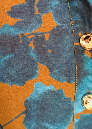 Close-up of a garment featuring a bold abstract floral pattern in teal blue and burnt orange, with a vertical seam and shiny gold buttons.